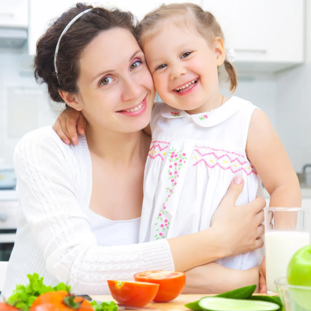 Smiling mother and daughter hugging in a bright kitchen with fresh vegetables on the table.